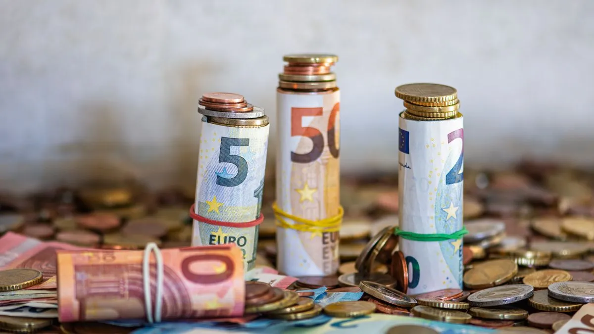 Rolled bundles of euro banknotes held with rubber bands, each topped with a stack of coins, standing on a spread of loose change.