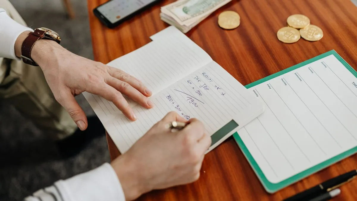 Notgroschen: Wie viele Monate brauchen Sie wirklich? A man's hands work through handwritten sums in a lined notebook, with folded banknotes, a few gold coins and a weekly planner sheet on the desk beside him.