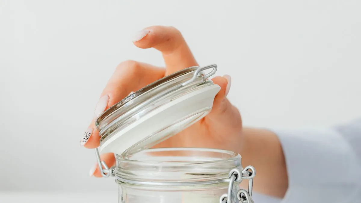 A woman's hand lifts the clip-top lid off an empty glass storage jar against a plain white background.