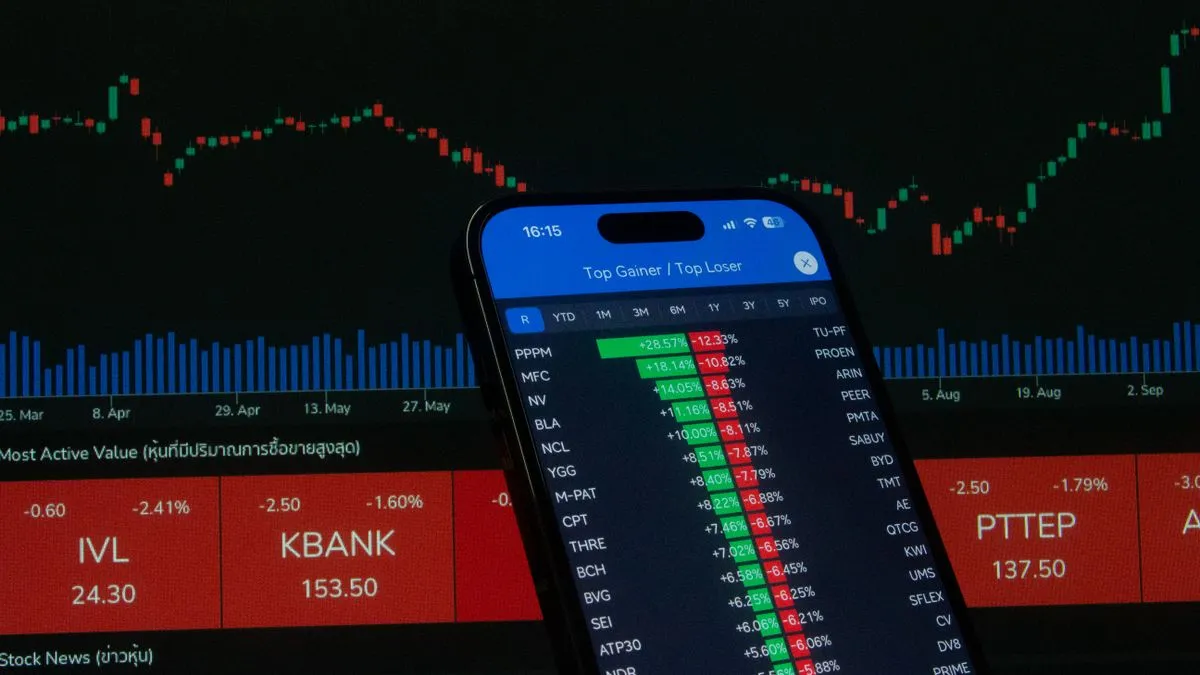A smartphone showing lists of green and red stock prices is held up in front of a large screen displaying market charts and tickers.