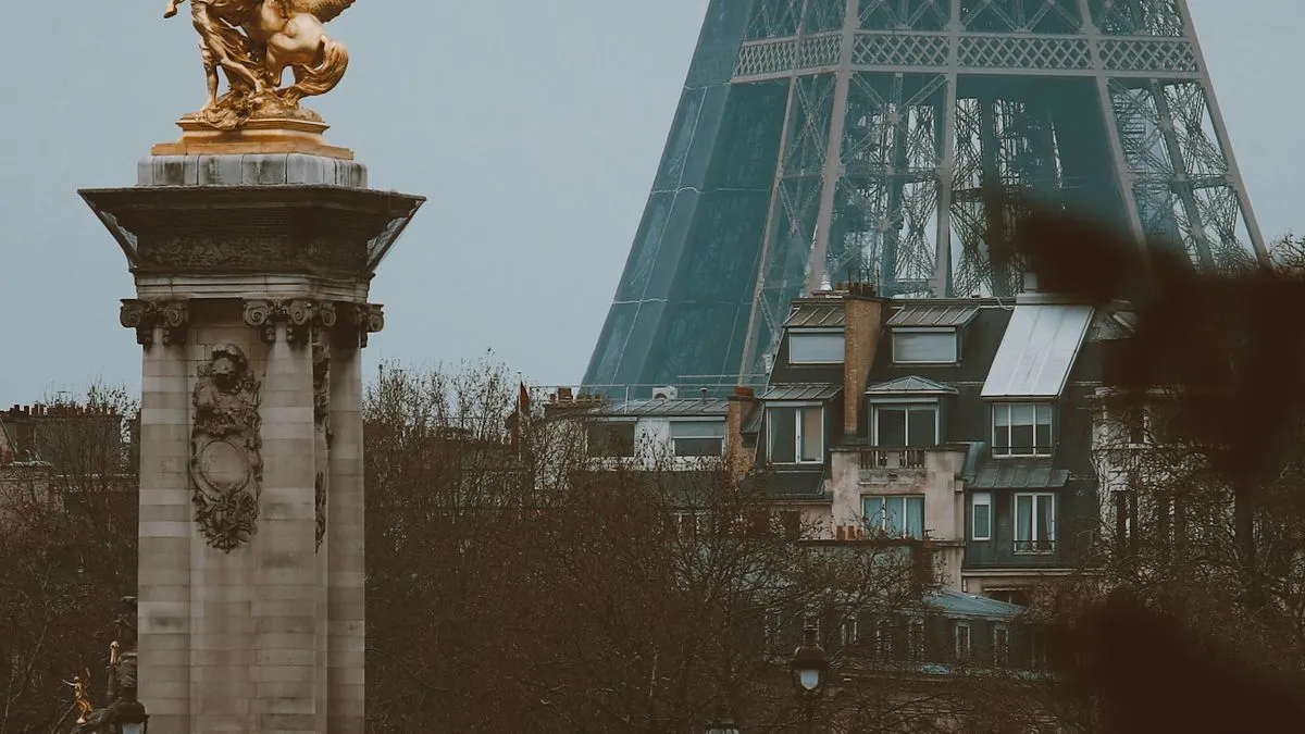 A gilded statue on a stone column stands before Parisian apartment buildings and the base of the Eiffel Tower.