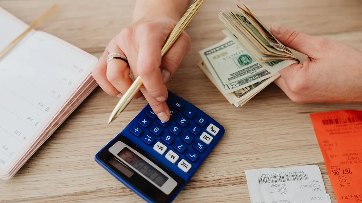 One hand taps a blue calculator with a pen while the other holds a fold of dollar bills over a desk.