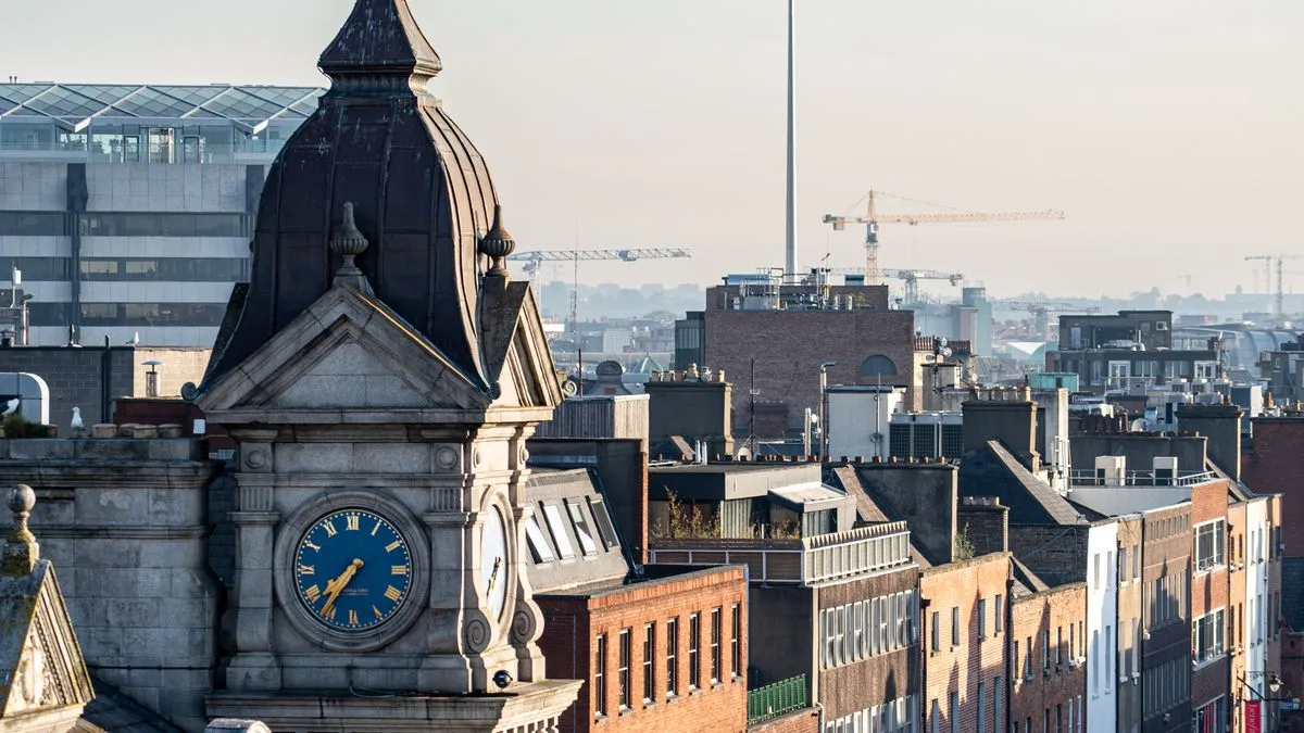 Ireland vs US Domiciled ETFs: What Actually Differs A view over Dublin rooftops, with an ornate stone clock tower in the foreground and construction cranes and the Spire on the hazy skyline.