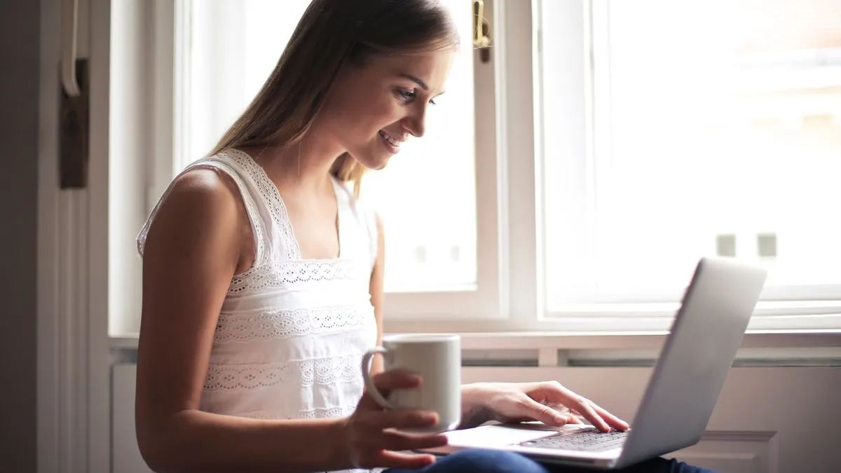 A smiling woman sits beside a bright window with a laptop on her lap and a white mug in one hand.
