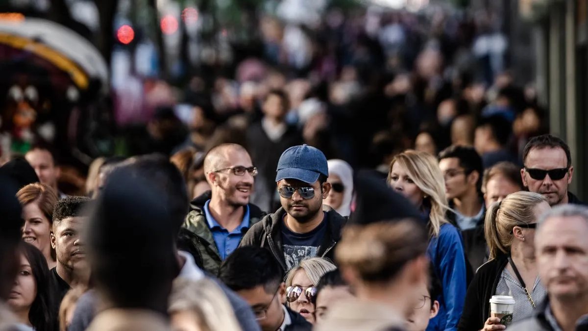 Nettovermögen nach Alter: Zahlen richtig einordnen A close-up of a crowded street, one man in a blue cap and sunglasses in sharp focus among dozens of blurred pedestrians.