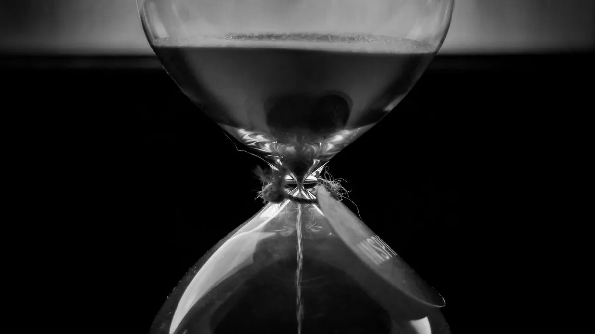 A black and white close-up of an hourglass, sand streaming through its narrow neck against a dark background.