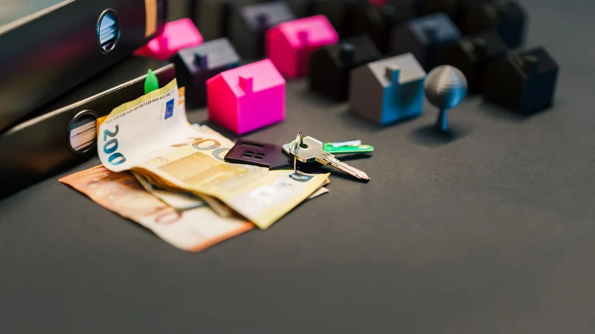Sondertilgung oder investieren? So entscheiden Sie Euro banknotes and a set of house keys lie on a dark desk beside a black folder, surrounded by small pink, grey and black model houses.