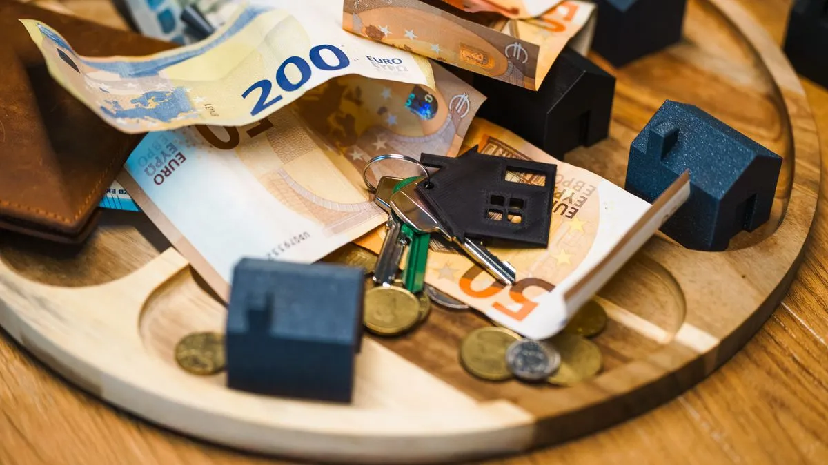 Euro banknotes, coins, house keys and small dark-blue model houses arranged together on a round wooden board.