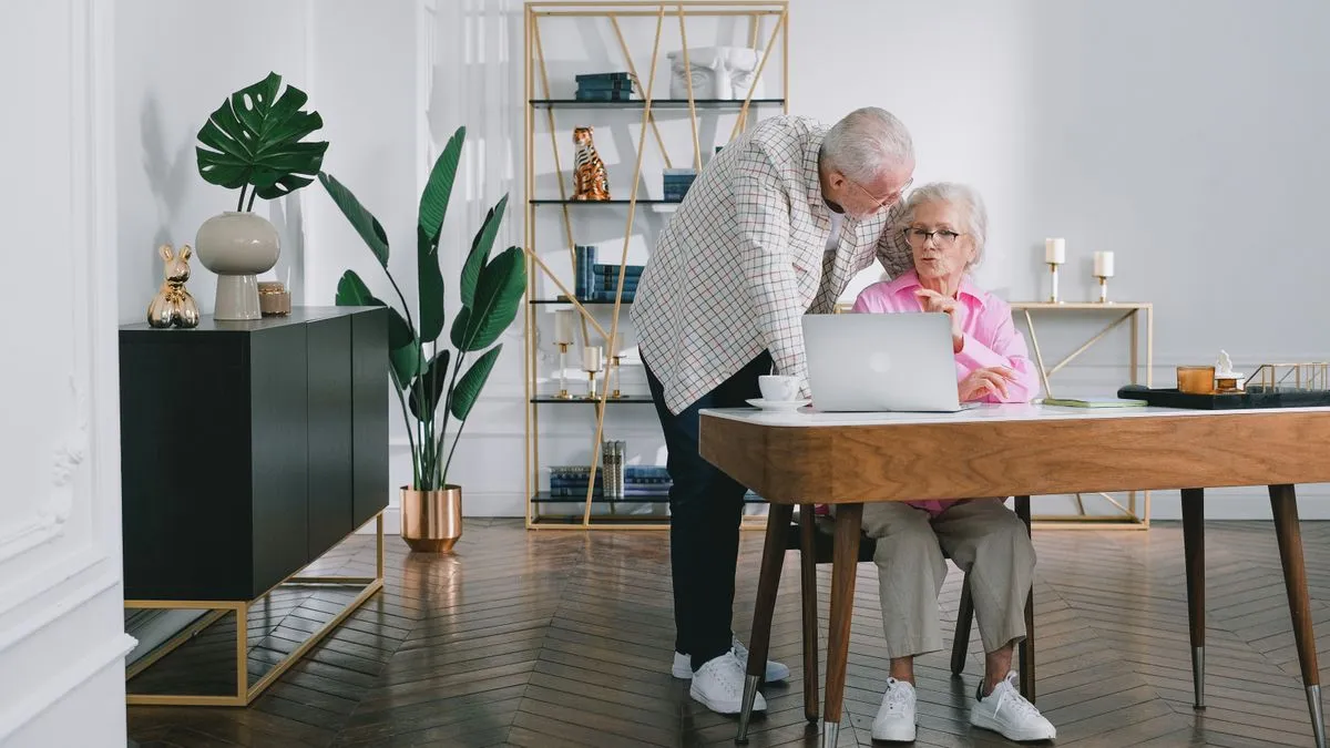 The Pension Gap: How to Size It Before You Fix It An older couple looks at a laptop together at a table at home, the man standing beside the seated woman.