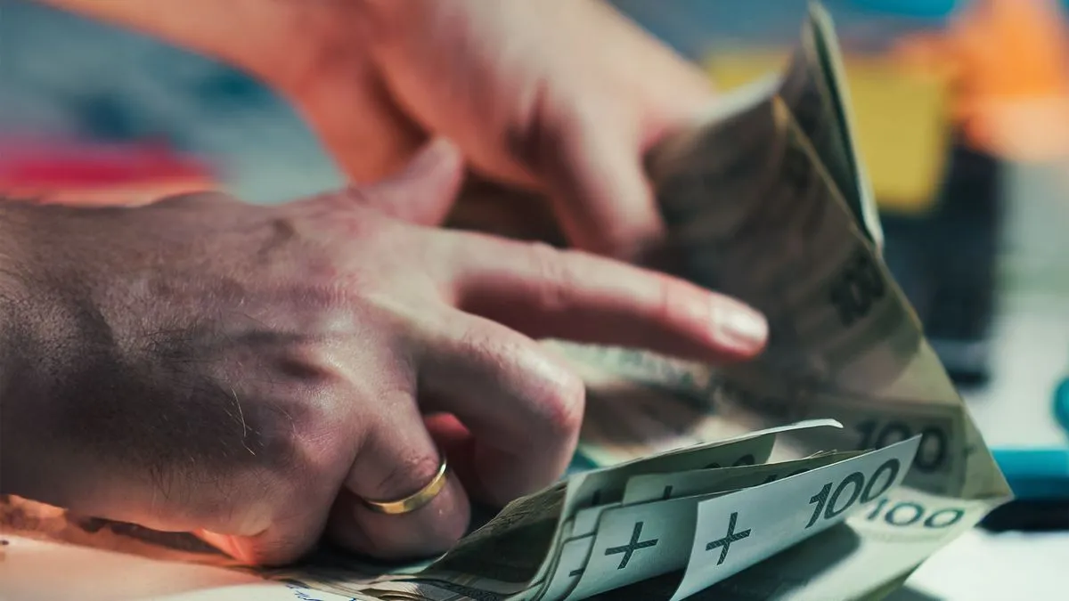 Close-up of a man's hands counting through a stack of banknotes marked 100 at a desk.
