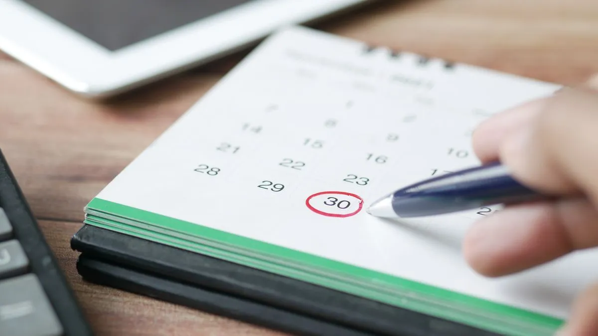 A hand with a pen marks a red circle around day 30 on a desk calendar beside a calculator.