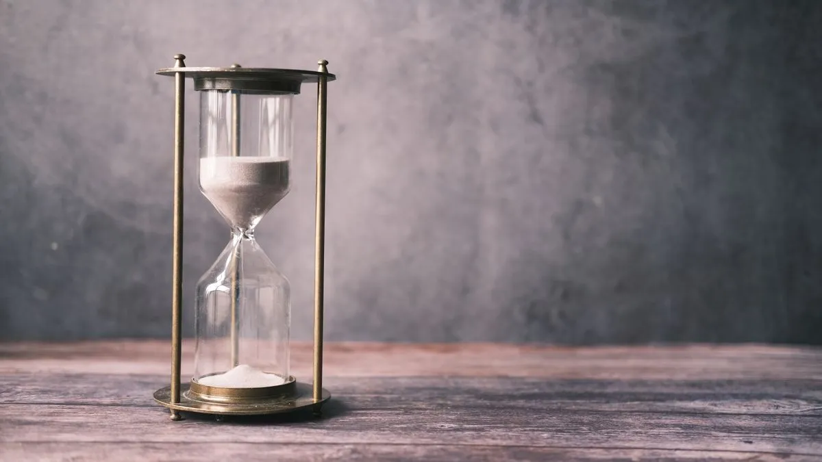 A brass-framed hourglass with white sand running sits on a wooden table against a grey wall.