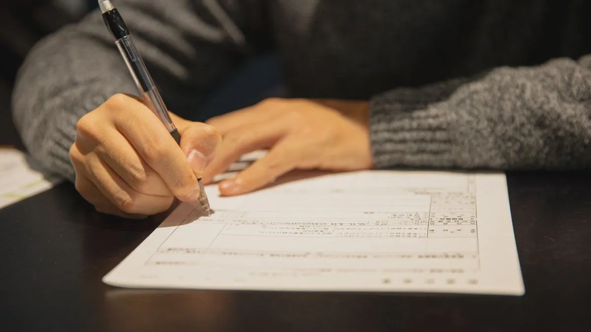 Freistellungsauftrag aufteilen: Sparerpauschbetrag nutzen A person in a grey sweater fills in a printed form with a ballpoint pen at a dark wooden table.