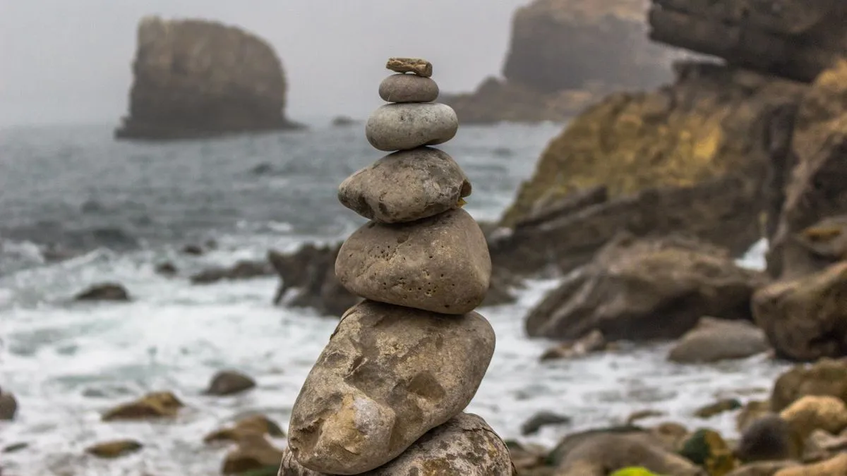 Weltportfolio mit 3 ETF: 70/30 richtig aufbauen A cairn of stacked stones stands on a rocky shoreline while waves break against misty cliffs behind it.