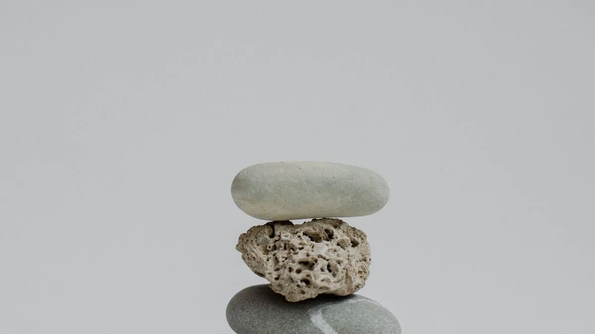 A small smooth pebble balances on a rough porous stone, which rests on a rounded grey base stone against a plain background.