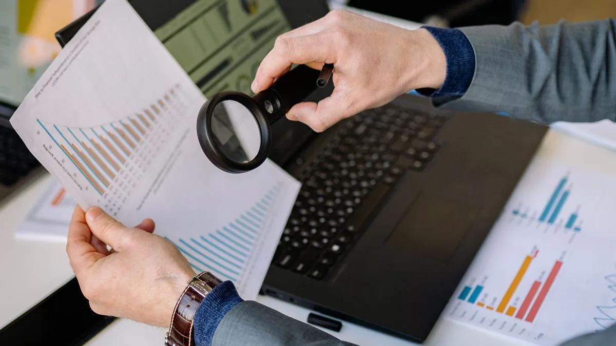 Aktien bewerten: KGV, KBV und was sie verschweigen Hands hold a magnifying glass over printed pages of bar charts and figures next to a laptop.