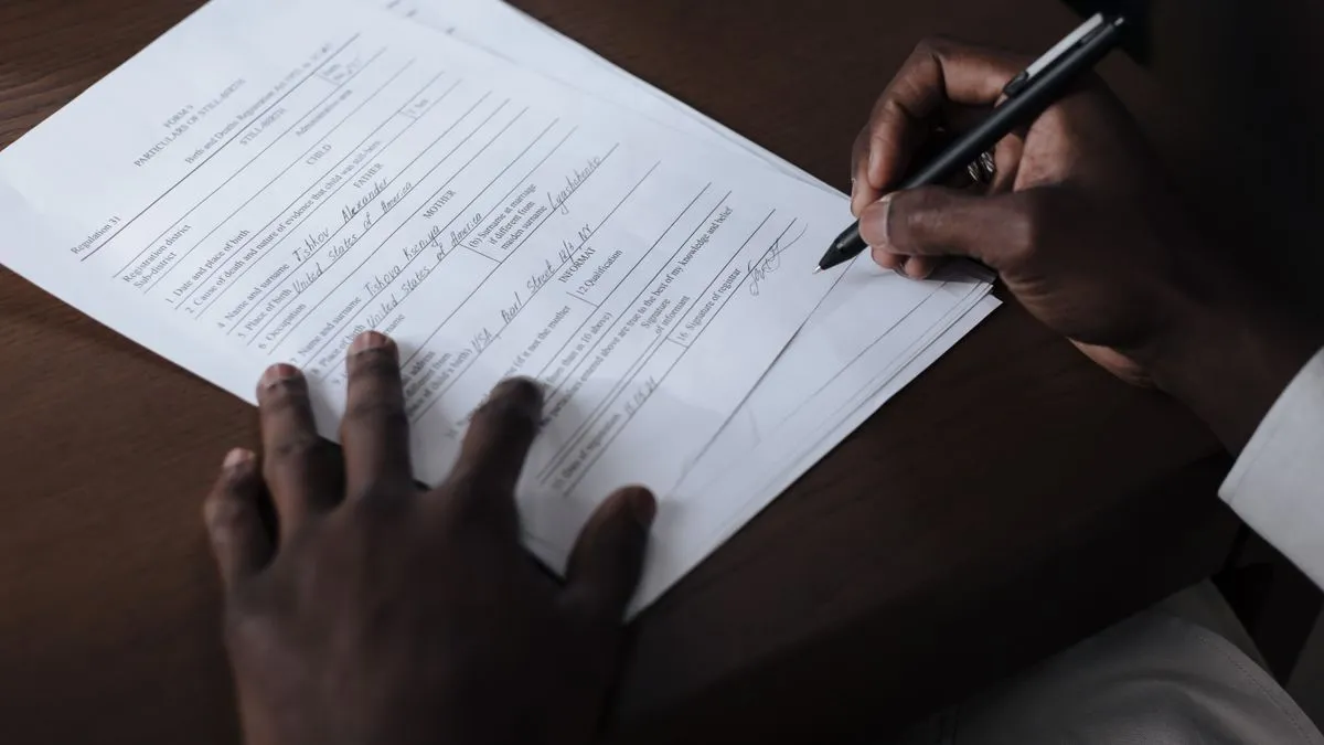 A close-up of two hands at a wooden desk, one steadying a printed form while the other signs it with a pen.