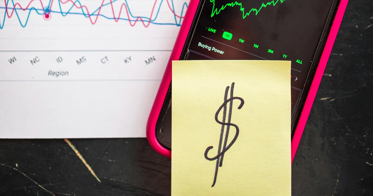 A phone showing a share price chart, next to a printed line chart and a sticky note with a dollar sign